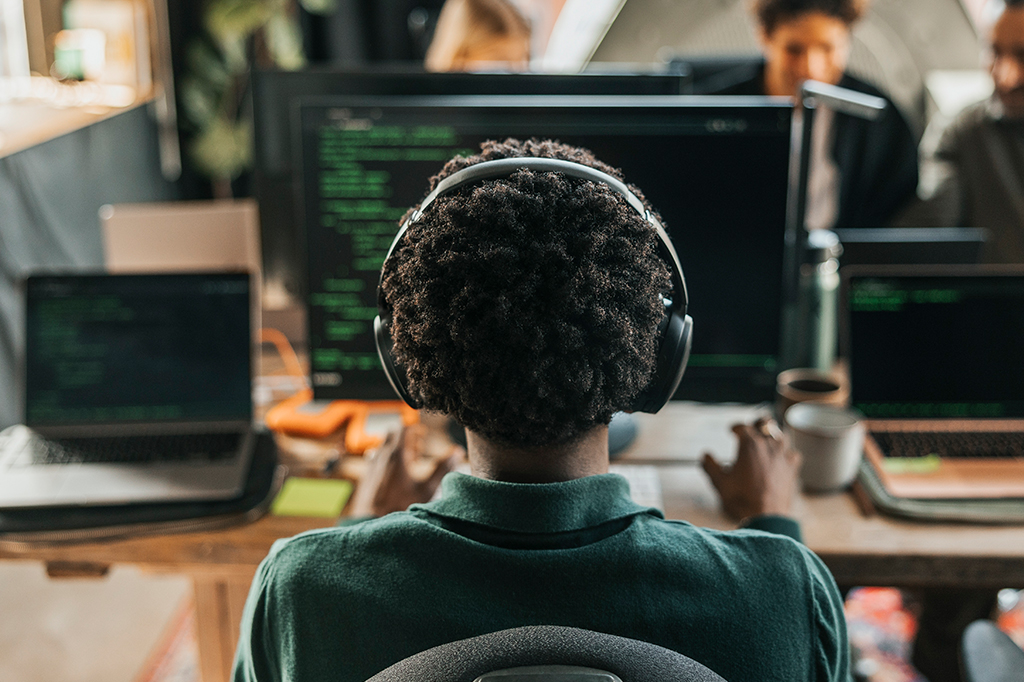 Pessoa de costas, com cabelo crespo e fones de ouvido, sentada em frente a um monitor exibindo códigos verdes. Ela usa uma camisa polo verde escura e digita no teclado. Outras pessoas e monitores desfocados aparecem ao fundo, em um ambiente de escritório.