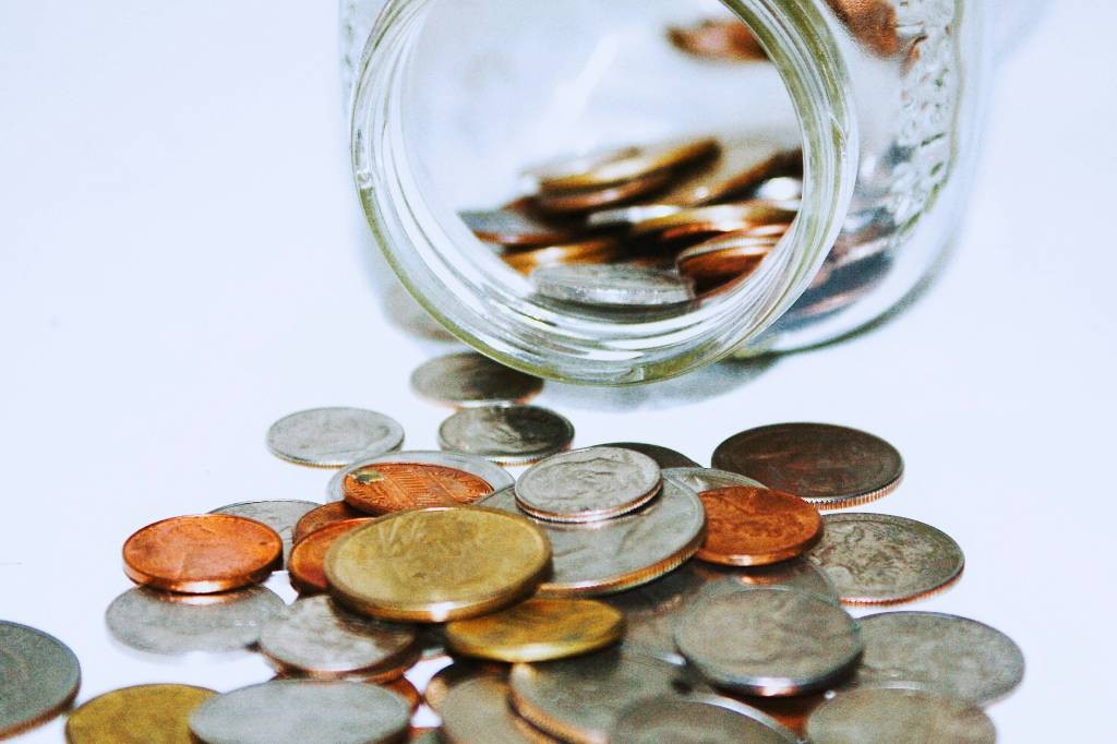 Um pote de vidro transparente tombado, derramando moedas de diferentes valores e cores, como prateadas, douradas e acobreadas, sobre uma superfície branca. As moedas estão espalhadas, algumas empilhadas, refletindo a luz