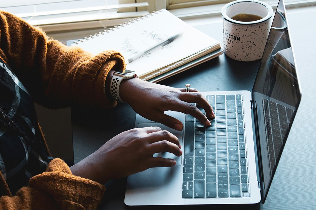 Mãos de uma pessoa com pele escura digitando em um laptop prateado, usando um suéter marrom e um relógio inteligente branco. Ao lado, um caderno, caneta e uma caneca branca com café.