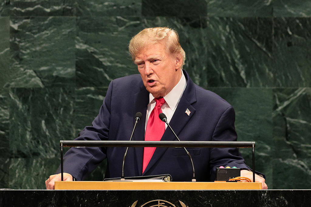 Donald Trump, com terno azul-marinho, camisa branca e gravata vermelha, discursa em um púlpito de madeira com microfones, contra um fundo de parede verde-escura. Ele tem a boca aberta, expressão séria e cabelos loiros. Uma pequena bandeira dos EUA está em sua lapela.