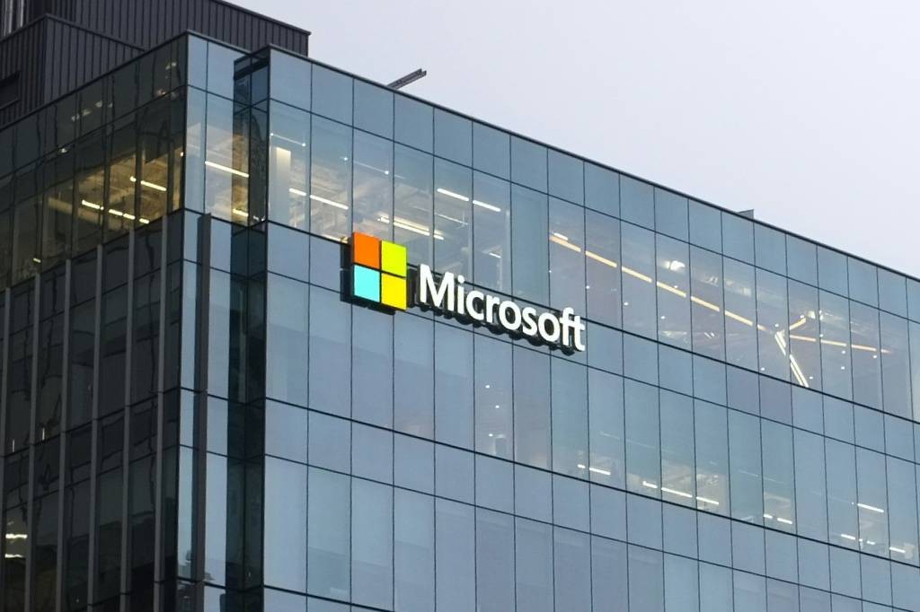 Edifício moderno com fachada de vidro espelhado, exibindo o logotipo da Microsoft em destaque. O logo tem quatro quadrados coloridos (laranja, verde, azul claro, amarelo) e o nome Microsoft em branco ao lado. O céu está nublado, e as luzes internas do prédio são visíveis através dos vidros