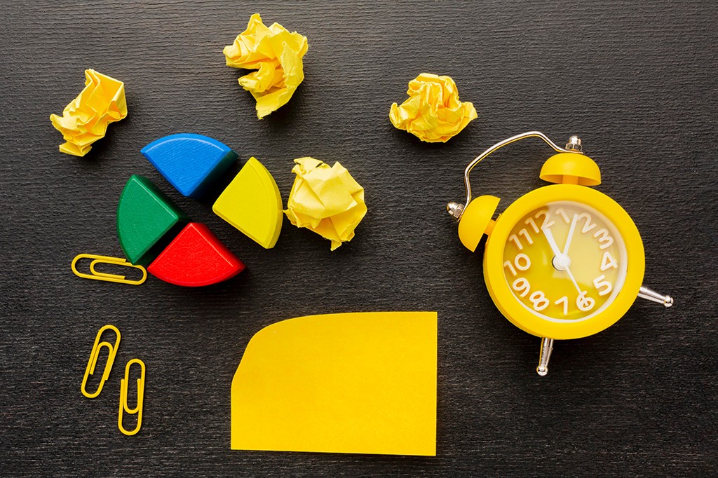 Objetos de escritório sobre uma superfície escura: um despertador amarelo, quatro pedaços de papel amassado, três clipes de papel e um bloco de notas amarelo, além de quatro peças coloridas de um gráfico de pizza (azul, verde, vermelho e amarelo)
